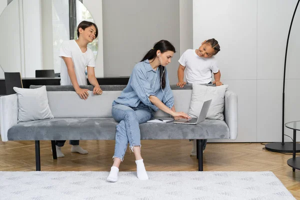 Mother in earphones using laptop near asian kids at home — Stock Photo