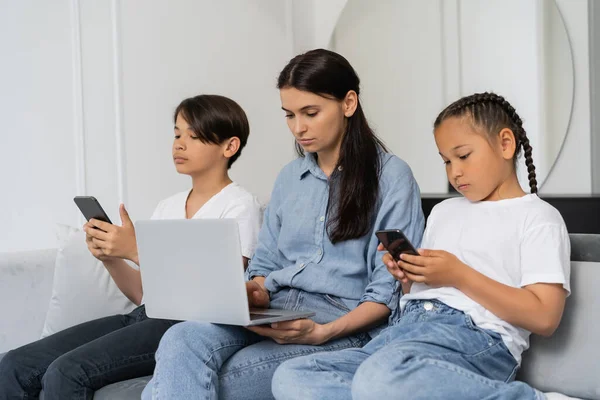 Bambini asiatici che utilizzano i telefoni cellulari mentre la madre lavora sul computer portatile a casa — Foto stock