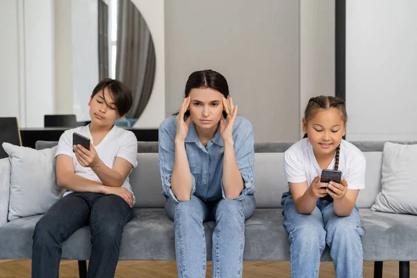 Donna che soffre di mal di testa vicino ai bambini asiatici utilizzando smartphone a casa — Foto stock