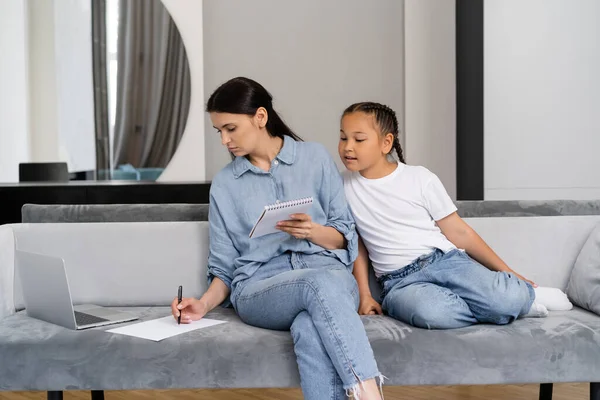 Woman writing on paper near laptop and asian daughter on couch at home — Stock Photo
