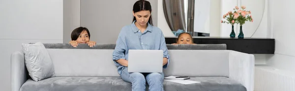 Sly asiático niños mirando mamá usando portátil en sofá en casa, bandera - foto de stock