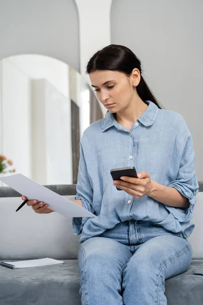 Femme brune tenant du papier et utilisant un smartphone à la maison — Photo de stock