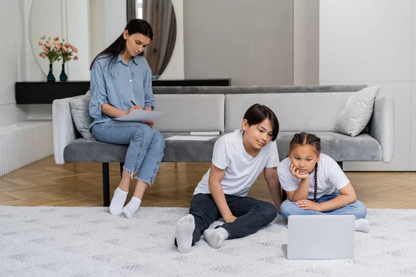 Asiatische Kinder schauen im Wohnzimmer auf Laptop neben Mutter, die auf Papier schreibt — Stockfoto