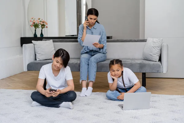 Asiatische Kinder benutzen Geräte, während die Mutter mit dem Smartphone spricht und Papier zu Hause hält — Stockfoto