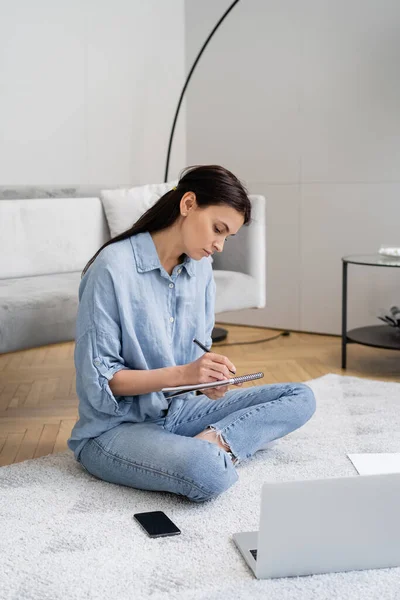 Brunette woman writing on notebook near devices on carpet at home — Stock Photo