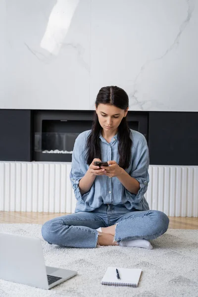 Brünette Freiberuflerin mit Smartphone in der Nähe verschwommenes Notebook und Laptop auf Teppich im Wohnzimmer — Stockfoto