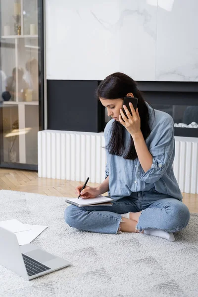 Freelancer talking on smartphone and writing on notebook near laptop and documents on floor at home — Stock Photo