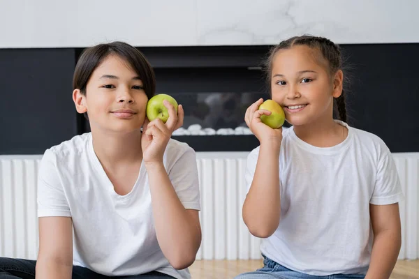 Lächelnde asiatische Geschwister, die zu Hause Äpfel halten und in die Kamera schauen — Stockfoto