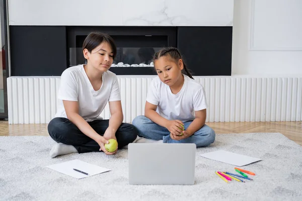 Bambini asiatici che tengono le mele e guardano film su laptop vicino alla carta e alle matite di colore a casa — Foto stock