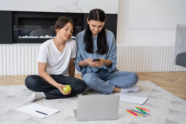 Mujer usando smartphone cerca asiático hijo con manzana mirando portátil en sala de estar - foto de stock