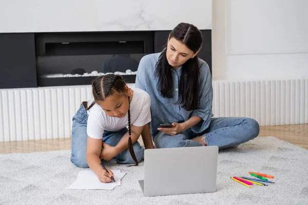 Menina asiática escrevendo no papel perto da mãe com smartphone e laptop em casa — Fotografia de Stock