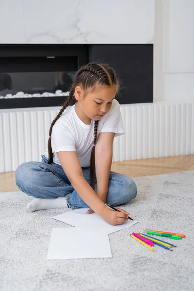 Niño asiático escribiendo en papel cerca de lápices de color en la alfombra en casa - foto de stock