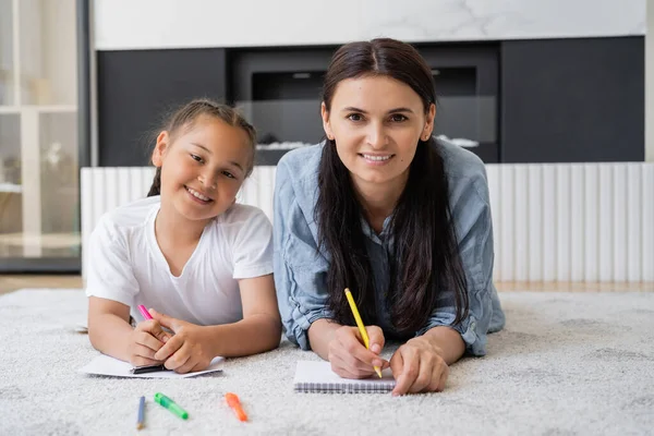 Sorridente donna che scrive su notebook e guardando la fotocamera vicino a figlia asiatica con marcatori a casa — Foto stock