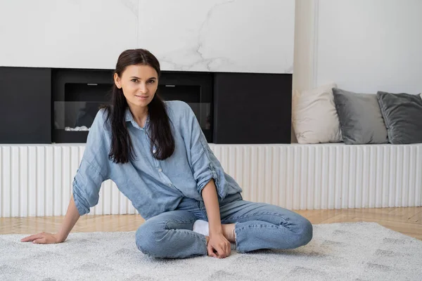 Brunette woman in shirt and jeans sitting on carpet in living room — Stock Photo