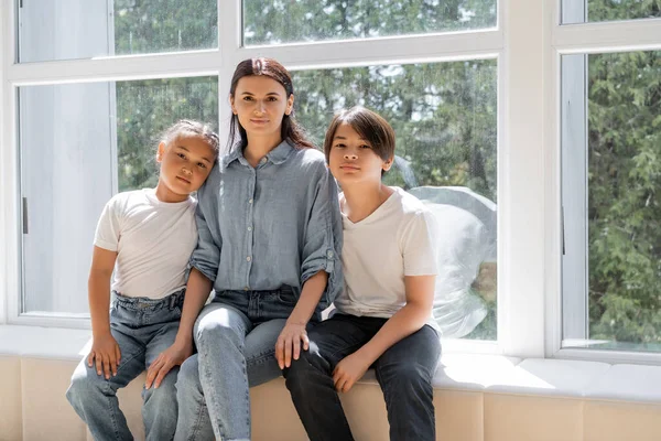 Femme et asiatique enfants regardant caméra près de fenêtre à la maison — Photo de stock