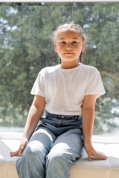 Portrait of asian girl looking at camera while sitting on windowsill at home — Stock Photo