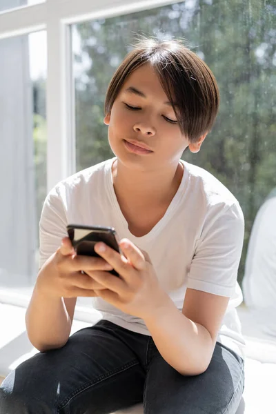 Preteen asian boy using smartphone while sitting on windowsill at home — Stock Photo