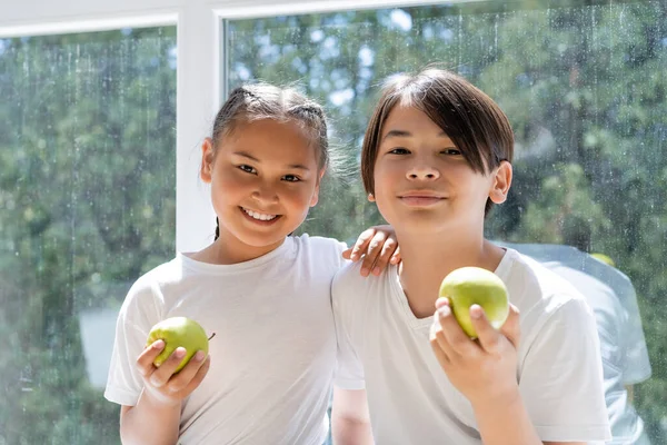 Smiling asian siblings holding fresh apples near window at home — Stock Photo