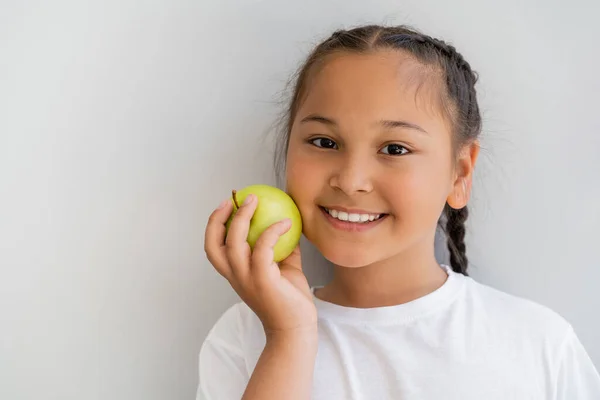Porträt eines lächelnden asiatischen Mädchens, das einen frischen Apfel in der Hand hält und in die Kamera neben der Wand blickt — Stockfoto