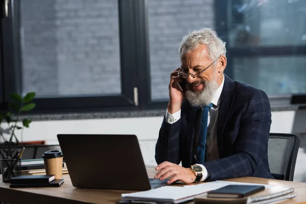 Cheerful mature businessman talking on cellphone near laptop and coffee to go in office in evening — Stock Photo