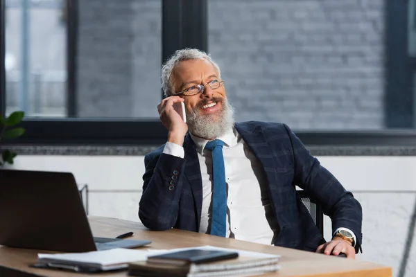 Empresário barbudo positivo em óculos falando no smartphone no escritório — Fotografia de Stock