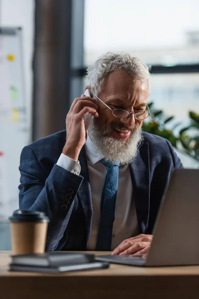 Positive reife Geschäftsmann im Anzug sprechen auf Smartphone in der Nähe von Laptop und Kaffee zum Mitnehmen im Büro — Stockfoto