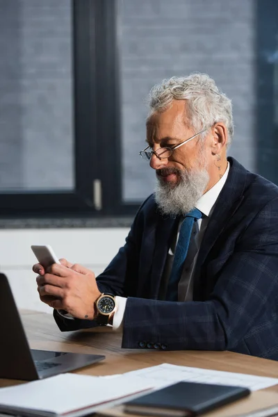 Bearded businessman in eyeglasses using cellphone near papers and laptop in office — Stock Photo