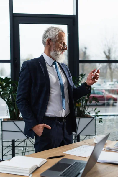 Side view of mature businessman in suit using smartphone near papers and coffee in office — Stock Photo