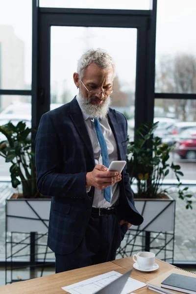 Smiling mature businessman in suit using smartphone near notebooks and coffee in office — Stock Photo