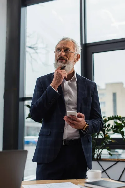 Pensive businessman holding smartphone and pen near coffee in office — Stock Photo