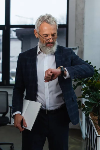 Mature businessman in suit looking at wristwatch and holding notebook in office — Stock Photo