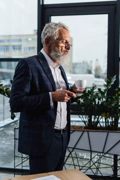 Middle aged businessman in eyeglasses holding cup of coffee in office — Stock Photo