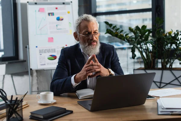 Uomo d'affari maturo in giacca e cravatta guardando laptop vicino a caffè e carte sul tavolo di lavoro — Foto stock