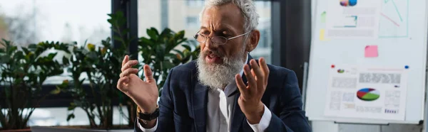 Middle aged businessman in eyeglasses gesturing near blurred flip chart in office, banner — Stock Photo