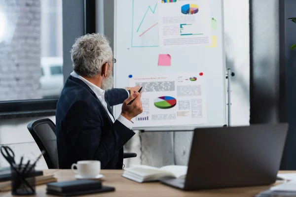 Grey haired businessman looking at blurred flip chart near laptop and coffee in office — Stock Photo