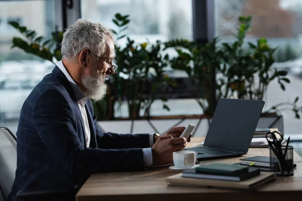 Side view of mature manager using devices near notebooks and coffee in office — Stock Photo