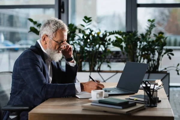Mature manager in eyeglasses writing on notebook near laptop and coffee in office — Stock Photo