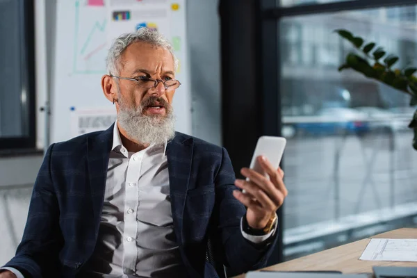 Mature manager in suit and eyeglasses using smartphone in office — Stock Photo