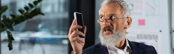 Middle aged businessman in eyeglasses holding smartphone and looking away in office, banner — Stock Photo