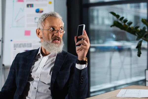 Grey haired businessman in eyeglasses holding smartphone and looking away in office — Stock Photo