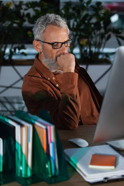 Концентрированный бизнесмен средних лет, рассматривающий компьютерный монитор вблизи размытых документов в офисе — стоковое фото