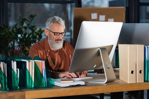 Empresario de mediana edad que usa computadora cerca del papeleo en la oficina por la noche - foto de stock