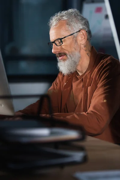 Татуйований бізнесмен в окулярах використовує комп'ютер в офісі ввечері — стокове фото