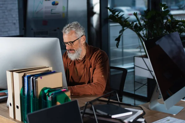Hombre de negocios de pelo gris en gafas que trabajan cerca de computadoras y documentos en la oficina - foto de stock