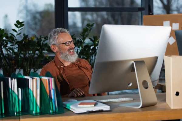Empresario positivo de mediana edad mirando hacia otro lado cerca de la computadora y los papeles en la oficina - foto de stock