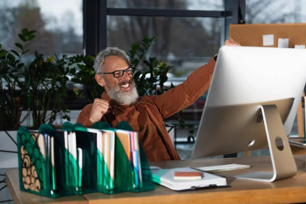 Emocionado hombre de negocios de pelo gris mostrando sí gesto cerca de la computadora y el papeleo en la oficina - foto de stock