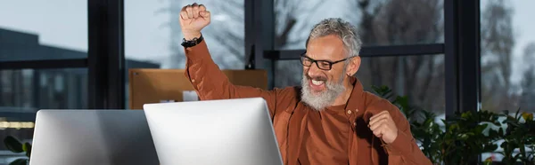 Middle aged businessman showing yes gesture near computer monitor in office in evening, banner — Stock Photo