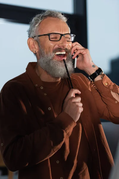 Allegro uomo d'affari barbuto con gli occhiali che tiene la matita mentre parla sullo smartphone in ufficio la sera — Foto stock