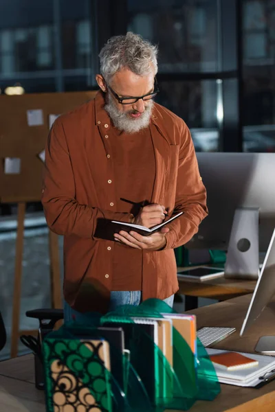 Middle aged manager writing on notebook near computers in office in evening — Stock Photo