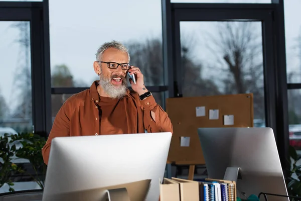 Позитивный бизнесмен в очках разговаривает на смартфоне рядом с компьютерами и бумажной работой в офисе — стоковое фото